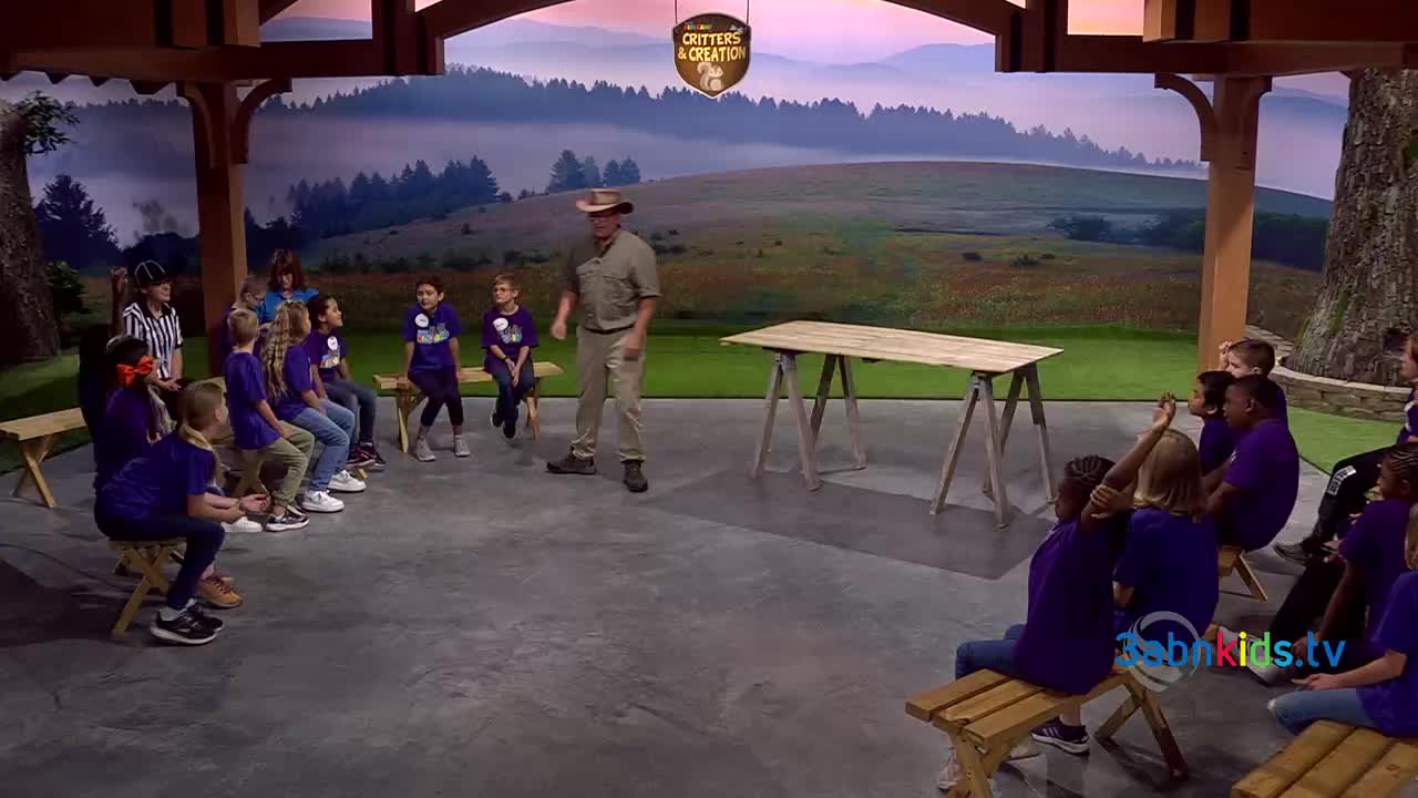 A man in a khaki outfit stands in front of a group of children, gesturing as if he's about to start a presentation. The kids, wearing purple shirts, are seated on benches and look towards him.
