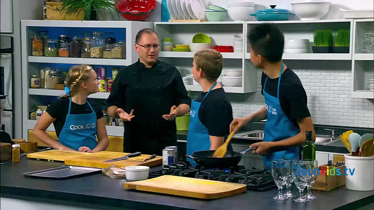 A chef in a black coat gestures as he speaks to three kids wearing aprons. One of the kids stirs something in a pan on the stove, while the others listen intently.
