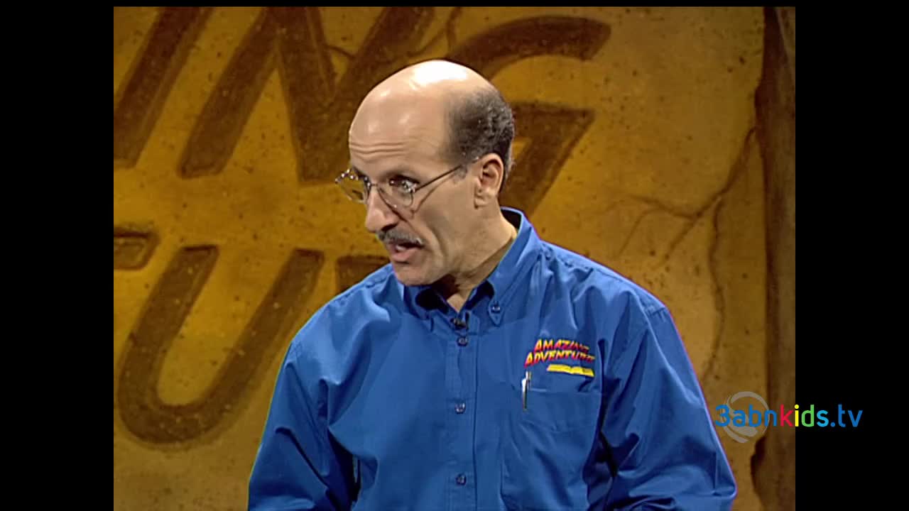 The man in the blue shirt is speaking, his mouth open as he looks to the side. Behind him, a textured wall with large, stylized lettering fills the background.
