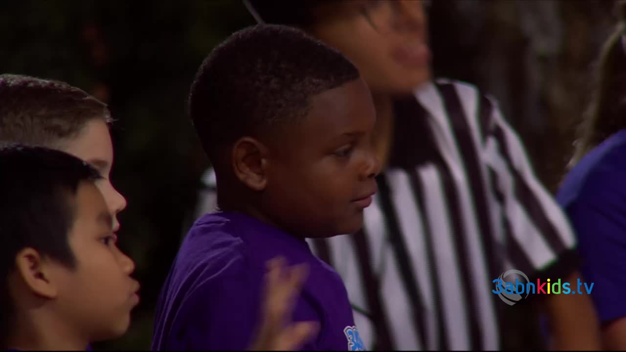 A young boy in a purple shirt looks to his right, his expression thoughtful. Behind him, a referee in black and white stripes stands ready.
