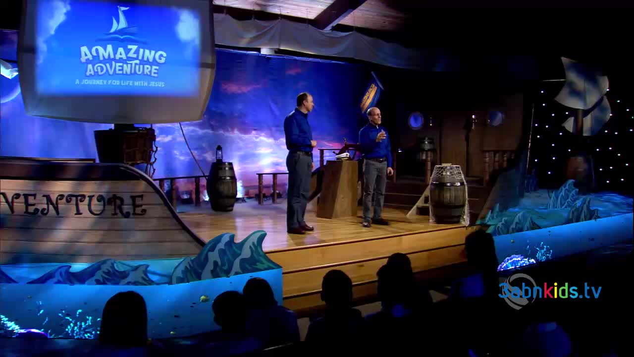 Two men in blue shirts stand on a stage designed to look like a ship, with a backdrop of a cloudy sky. The words "Amazing Adventure" are displayed on a screen above them.
