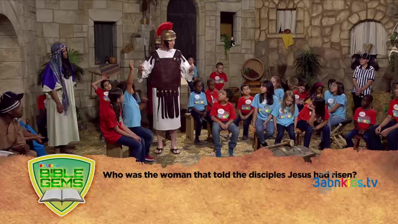 A man in a Roman soldier costume gestures as he speaks to a group of children. Several kids in matching shirts are seated, and one has raised their hand to answer a question.
