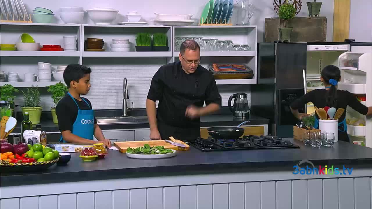 A man in a chef's coat gestures toward a pan on the stove, while a young boy in an apron watches. A girl opens the refrigerator, reaching inside for something.
