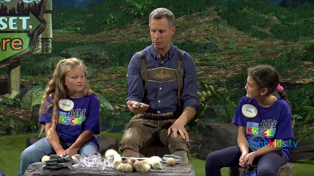 A man in leather suspenders holds small stones, speaking to two girls wearing "3ABN Kids Camp" shirts. They're seated on a log in a set designed to look like a forest.
