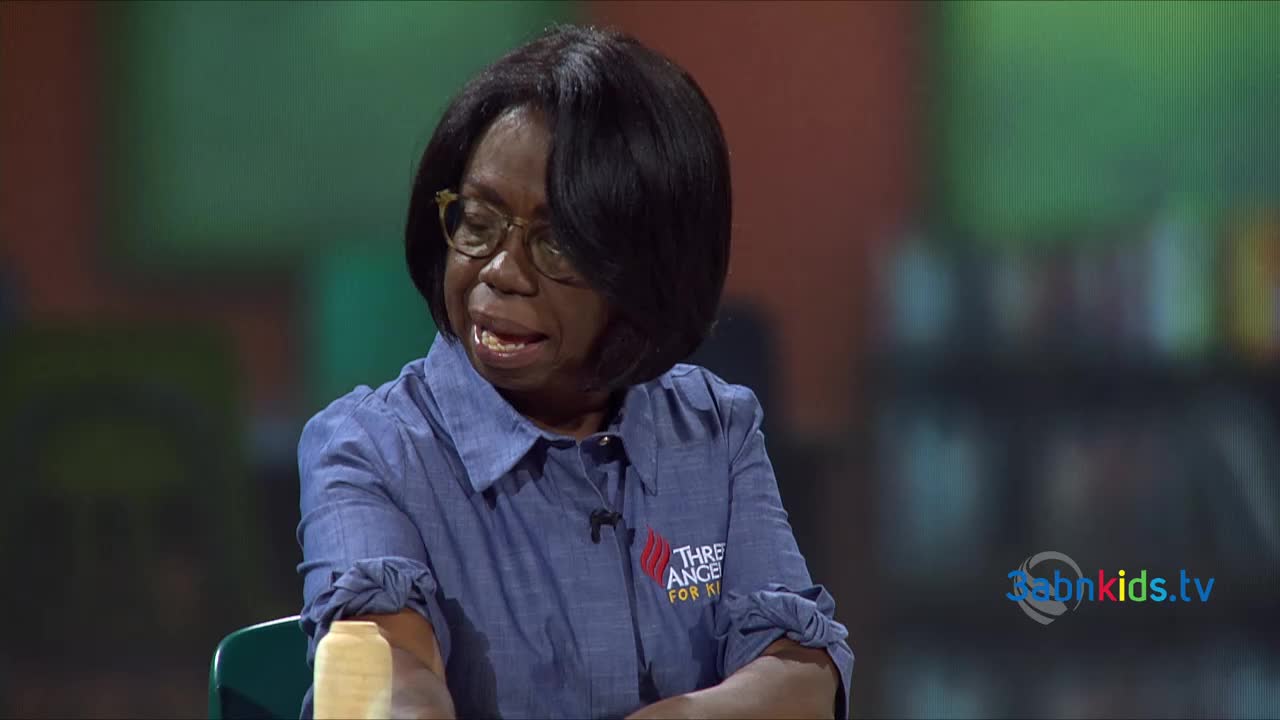 A woman in glasses and a blue shirt is speaking, her mouth open as if mid-sentence. The scene is set on a stage, likely for a program from the United States, 3ABN Kids.

