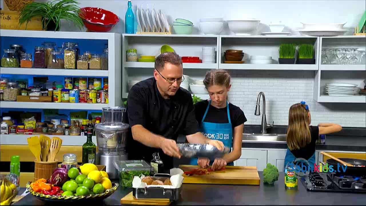 A man in a black chef's coat and a girl in an apron are working together, preparing food in a bright kitchen. A younger girl is standing nearby, focused on a pan on the stove.
