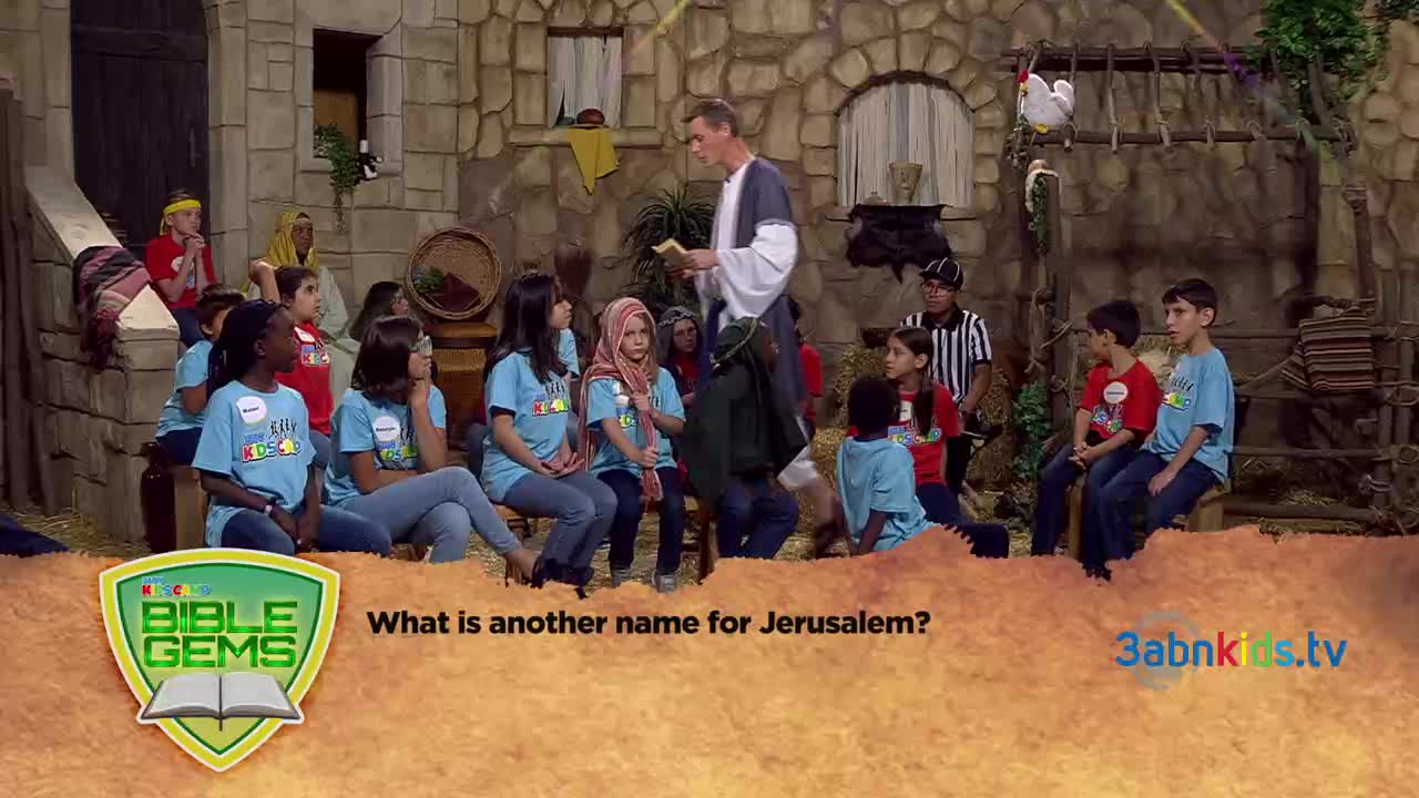 A man in a robe reads from a book in front of a group of children, all on a set designed to look like a stone building. The 3ABN Kids show is in progress, with a question about Jerusalem displayed on the screen.
