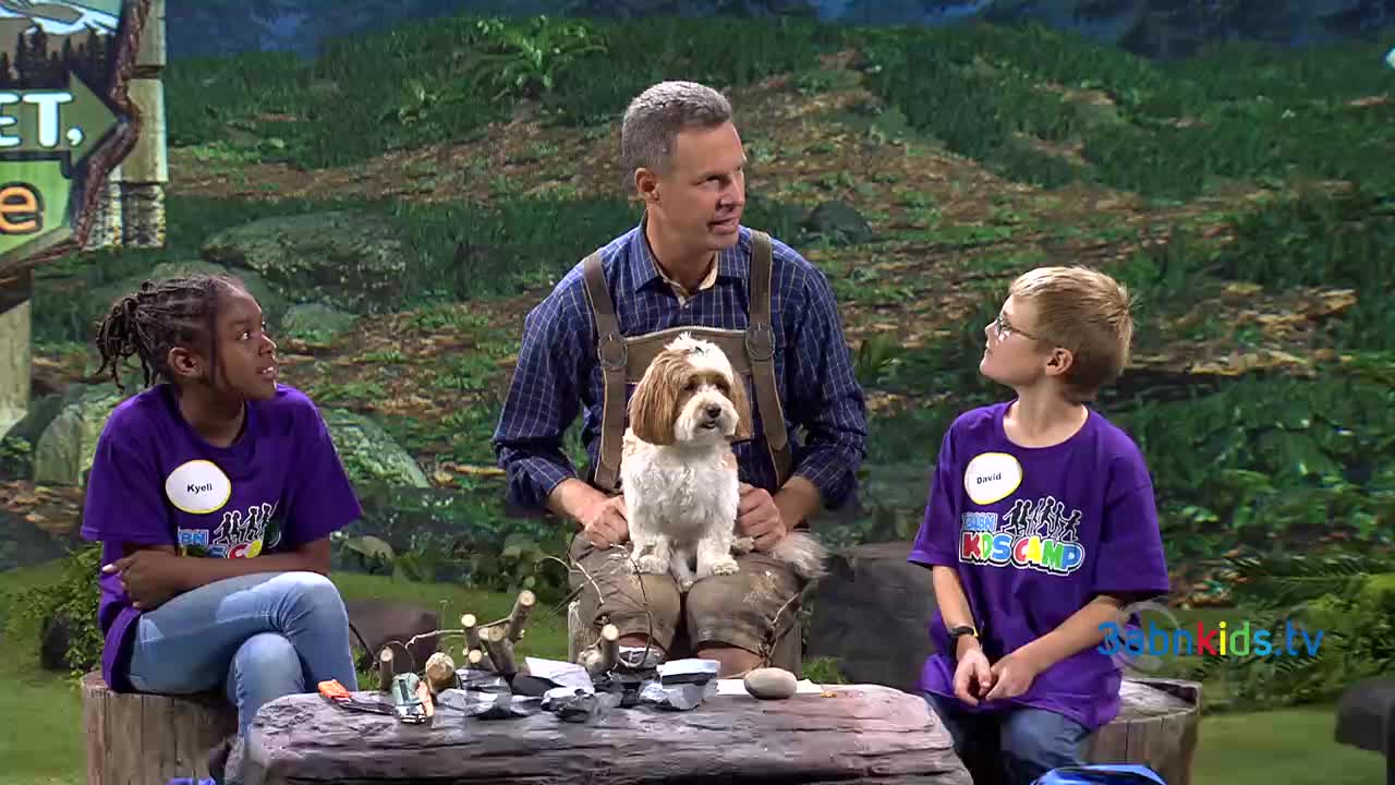 A man in overalls holds a fluffy dog on his lap, flanked by two children wearing matching purple shirts. They're all gathered around a table with a camping theme, likely on a set for 3ABN Kids.
