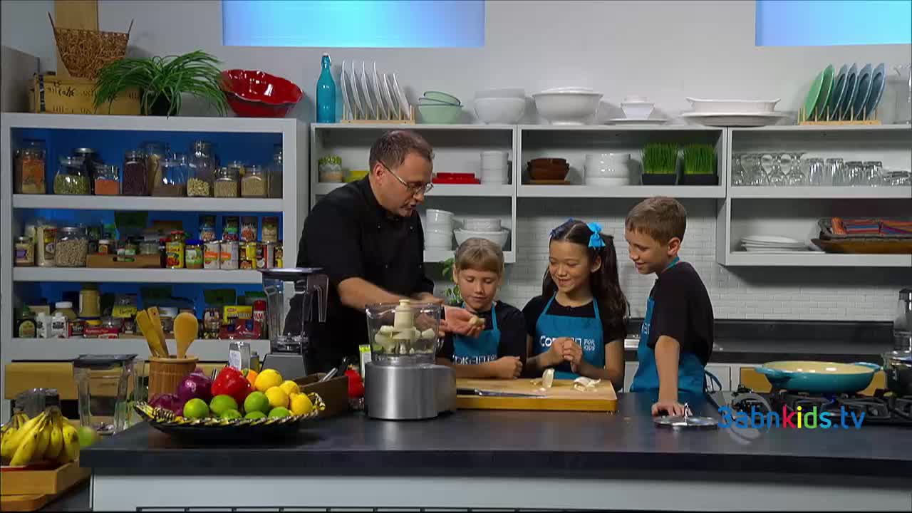 A man in a black chef's coat shows three children how to put something into a food processor. They're all gathered around a counter in a kitchen studio, likely filming an episode of 3ABN Kids.
