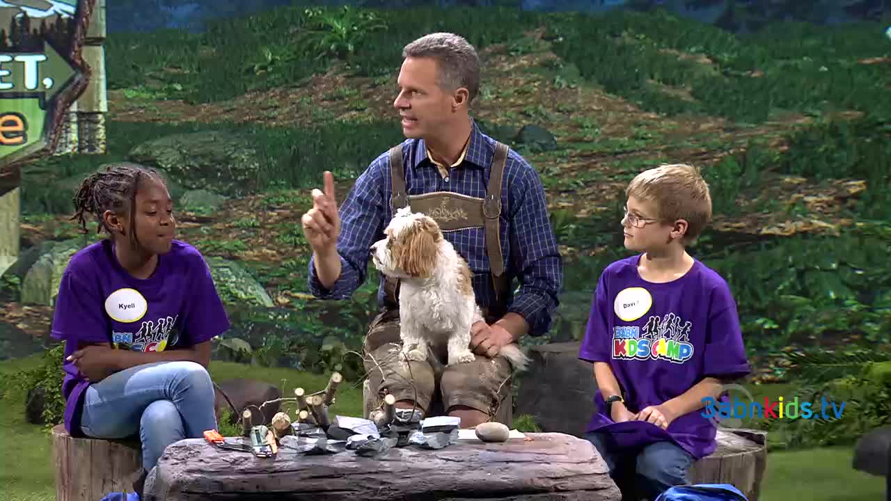 A man in lederhosen gestures while a small dog sits on his lap. Two children in matching purple shirts listen intently on either side of him.

