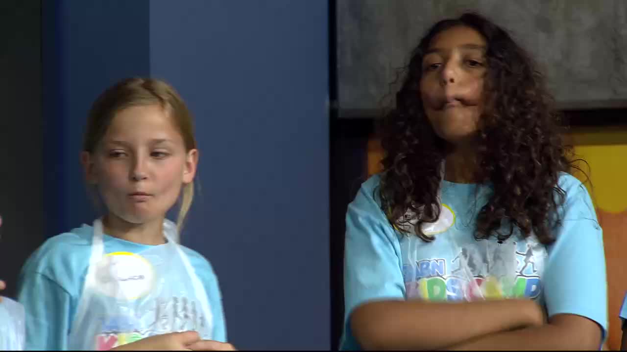 Two girls in light blue t-shirts stand side-by-side. The girl on the left, with blonde hair, looks to her right with her mouth slightly open. The girl on the right, with curly dark hair, has her arms crossed and her lips pursed.