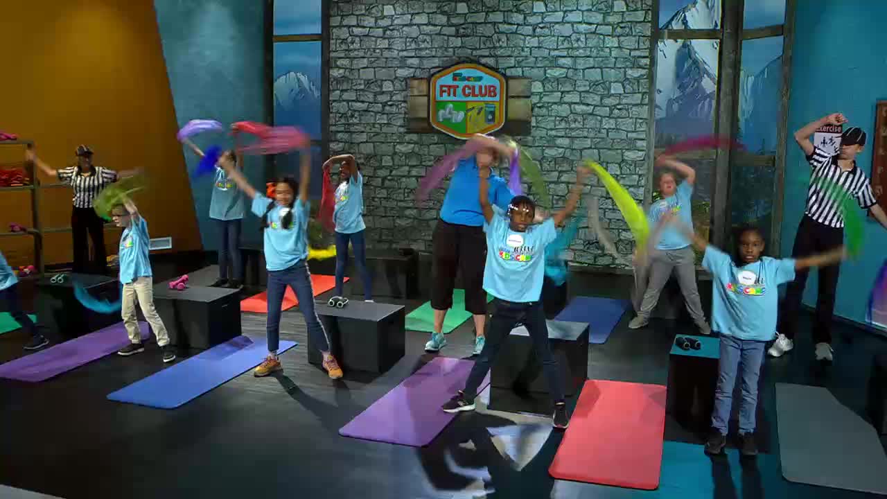 Children in blue shirts are waving colorful scarves overhead. A woman in a blue shirt leads them in an exercise routine.