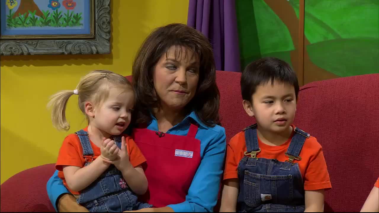 A woman with dark hair sits on a red couch, holding a young girl with blonde pigtails on her lap. To her right, a young boy in denim overalls looks off into the distance.