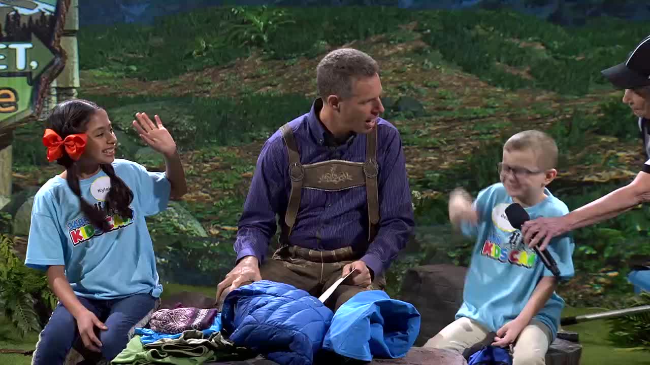 A young girl in a bright blue shirt with "3ABN Kids" on it waves enthusiastically. Beside her, a boy in a similar shirt holds a microphone and gestures with his hand.