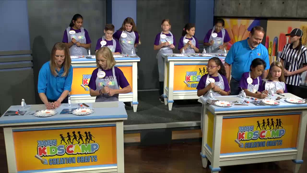 A group of kids are busy with crafts at their tables, some wearing purple shirts and aprons. A woman in a blue shirt guides one of the children, while a man in a blue polo and a woman in a referee shirt stand nearby.