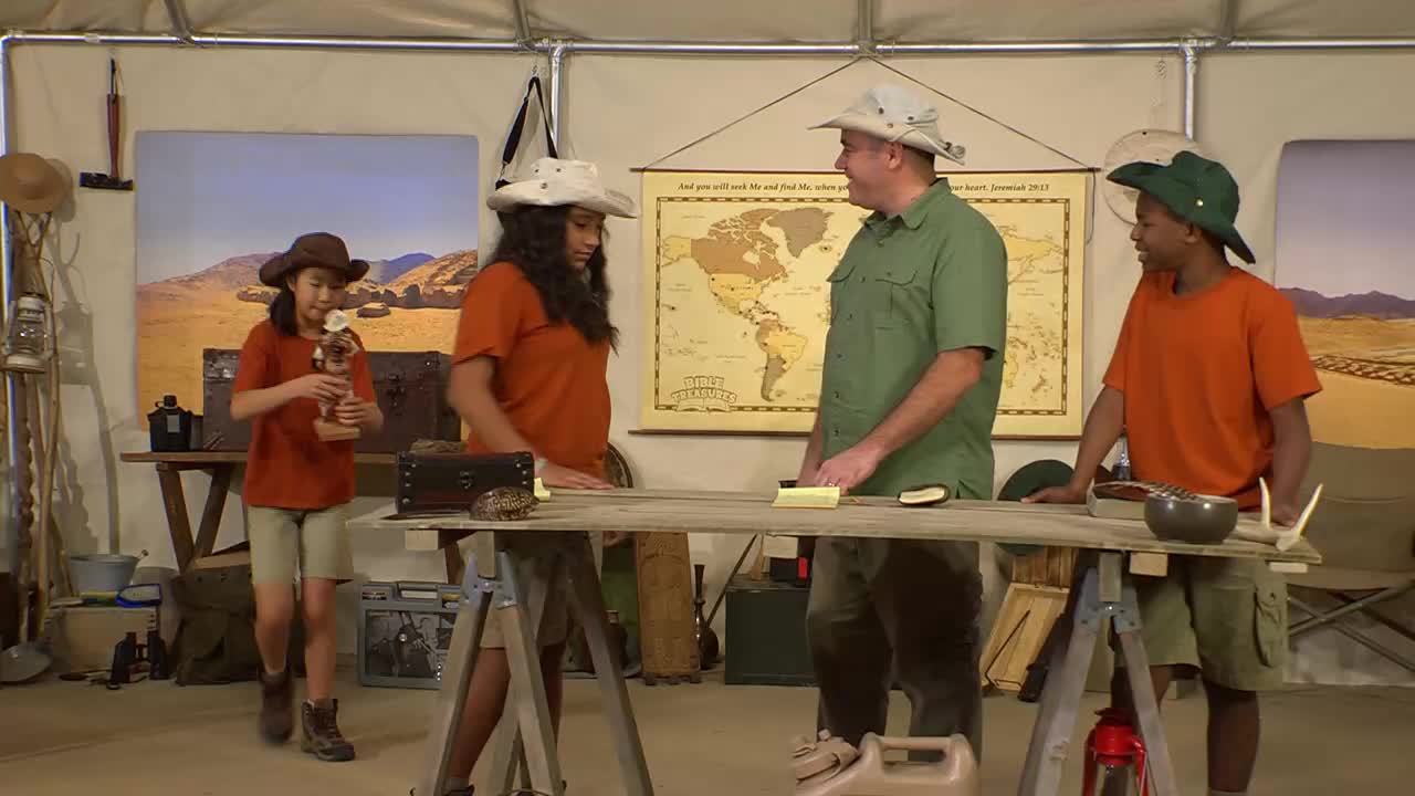 A young girl in a brown hat walks forward, holding a wooden trophy. A man in a green shirt and two kids in orange shirts stand around a table.