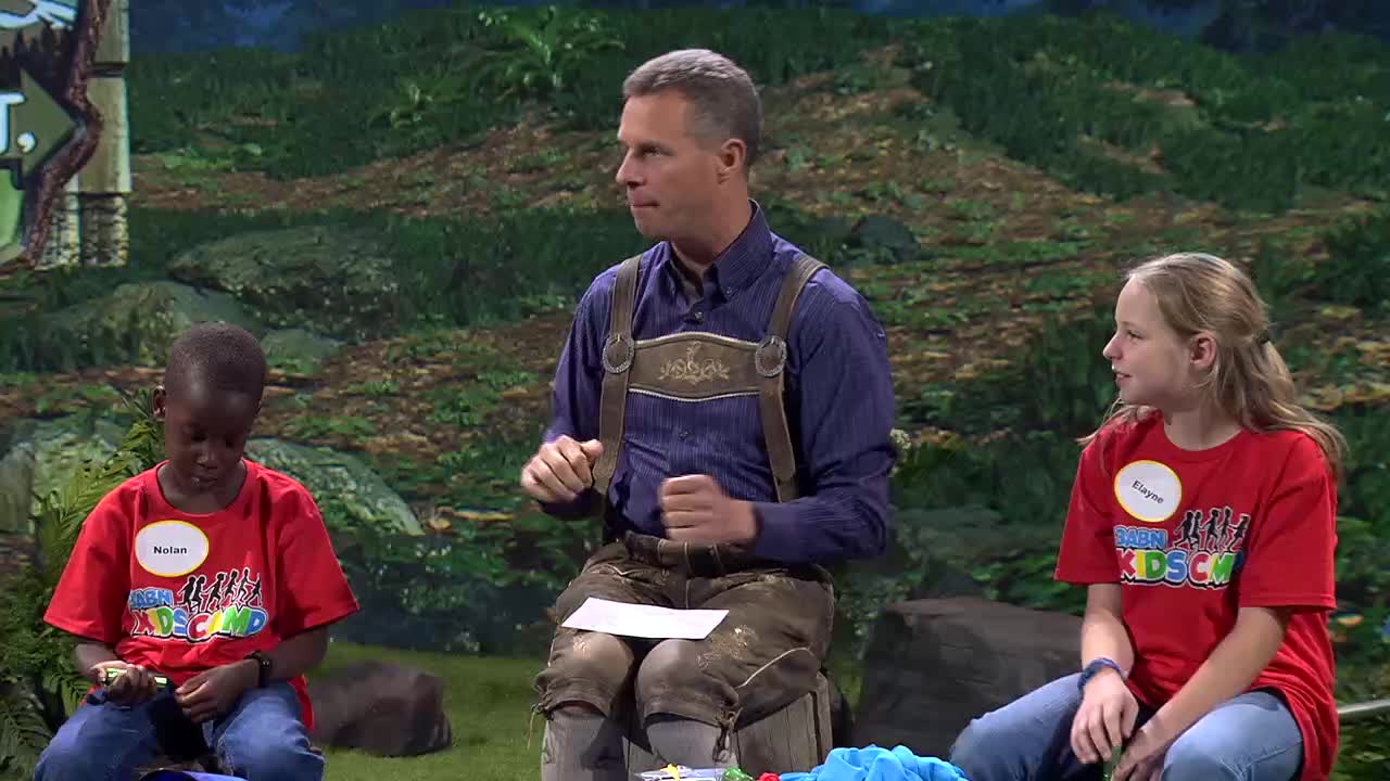 A man in lederhosen gestures with his hands while speaking to two children. The boy on the left, Nolan, looks down at something in his lap, and the girl on the right, Elayne, faces forward. They are all wearing red shirts with "3ABN Kids" printed on them.