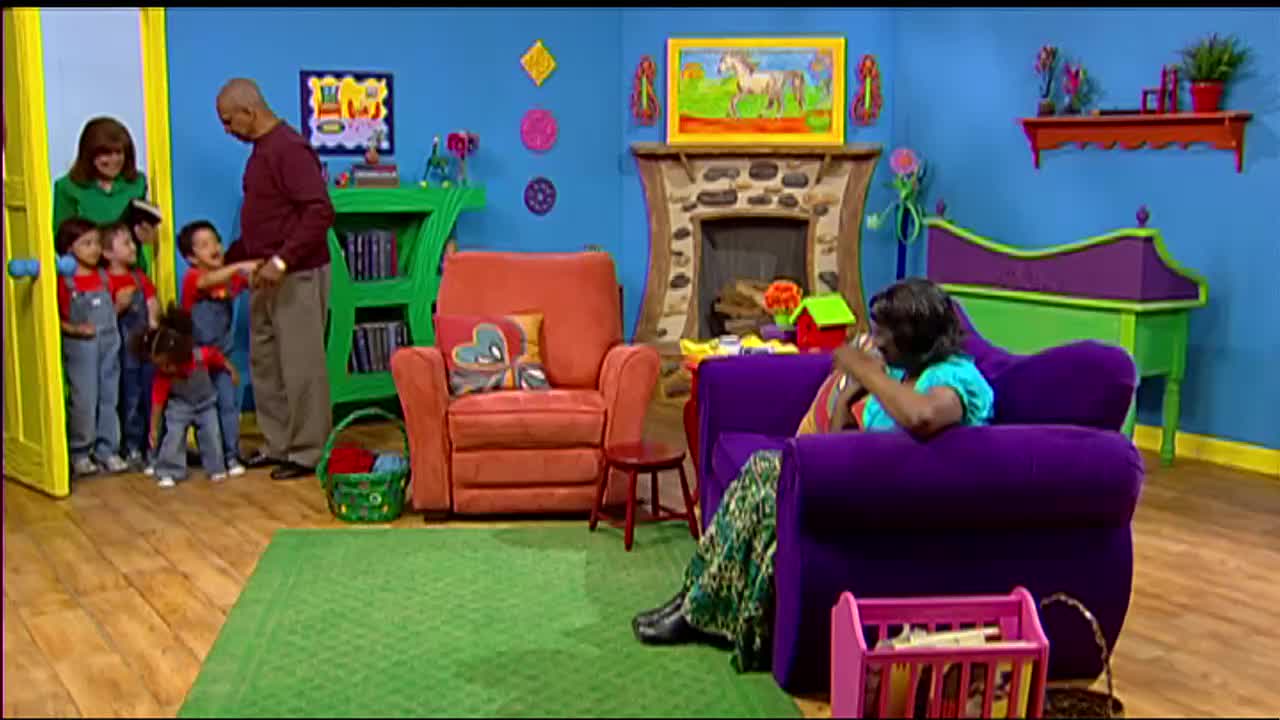 A woman in a green shirt holds a book, ushering a group of children in red shirts and blue overalls through a yellow doorway. A man in a maroon shirt stands nearby, watching them enter the colorful room.