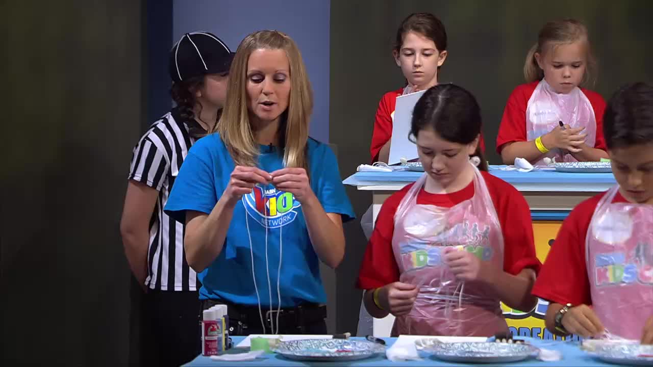 A woman in a blue shirt demonstrates a craft to children wearing red shirts and plastic aprons. A person in a referee's uniform stands behind her.