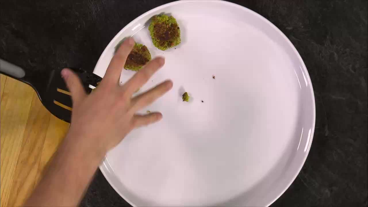 A hand reaches out to arrange two small, green patties on a white plate. A slotted spatula rests on the edge of the plate, ready to help.