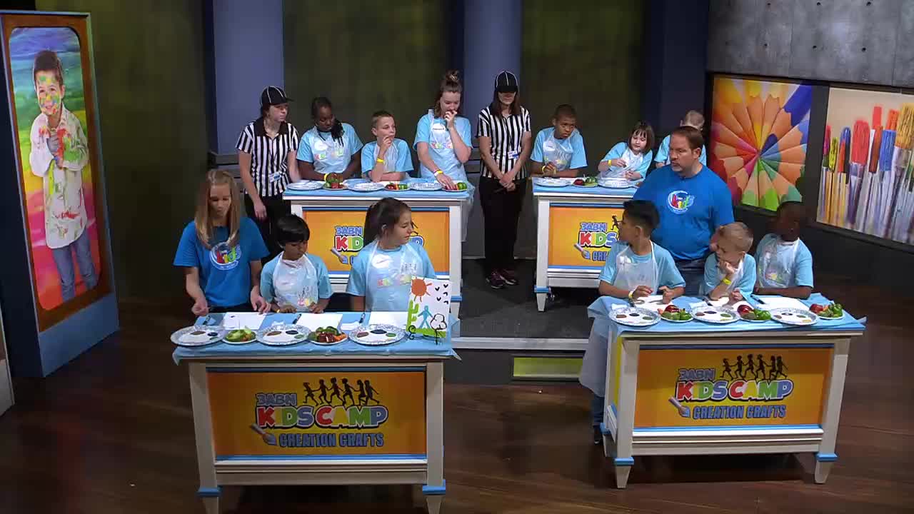 Kids in light blue shirts and white aprons stand at tables, preparing food for a competition. A man in a bright blue shirt watches them, while others in striped shirts stand behind them.