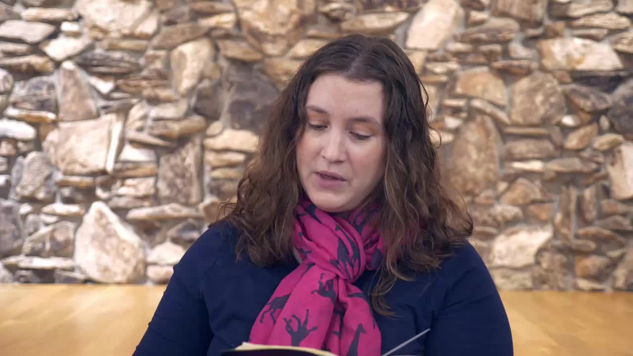 A woman with long, wavy brown hair, wearing a navy blue shirt and a bright pink scarf with a giraffe pattern, reads from a book. She's seated in front of a textured stone wall.
