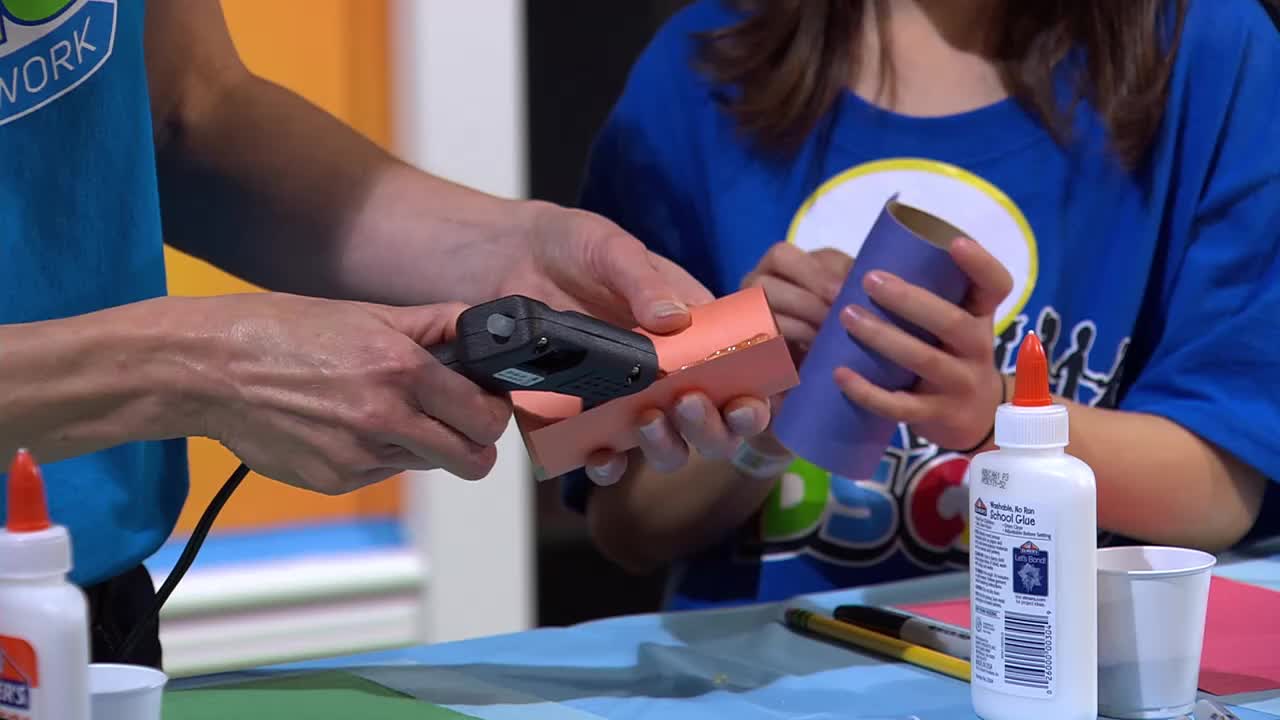 Hands hold a glue gun to an orange paper tube, while another child clutches a blue one. A bottle of Elmer's glue stands ready on the table.