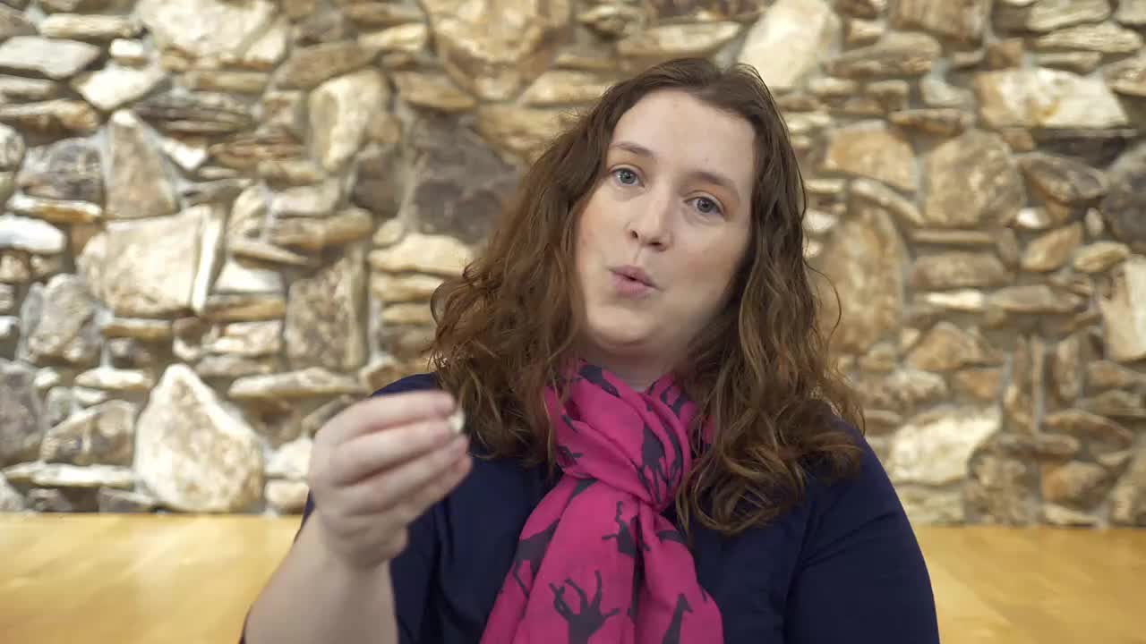 A woman with curly brown hair, wearing a navy shirt and a pink scarf, holds a small, light-colored object between her thumb and forefinger. She speaks directly to the camera, her mouth slightly open, against a backdrop of stacked, rough-hewn stones.