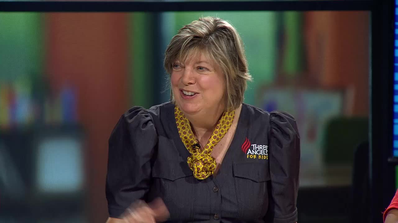 A woman with a bright smile is speaking, her hands gesturing animatedly. Her dark shirt features a small logo for "Three Angels for Kids," a program from the United States.