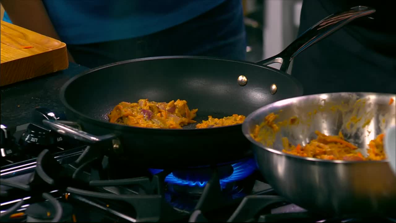 A bright blue flame heats a black pan where a mixture of orange and red ingredients is cooking. Next to it, a metal bowl holds more of the same mixture.