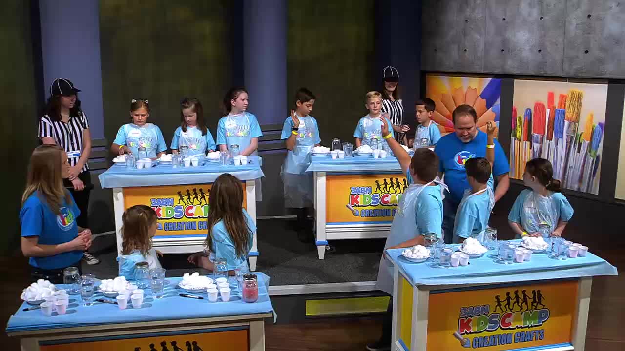 A man in a blue shirt guides children in a science experiment for 3ABN Kids. Several kids, wearing light blue shirts, stand behind tables laden with small cups and white powder.