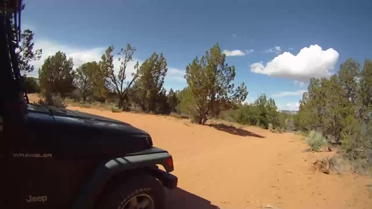 The Jeep Wrangler climbs a sandy incline under a bright blue sky. Scrubby trees dot the desert landscape.