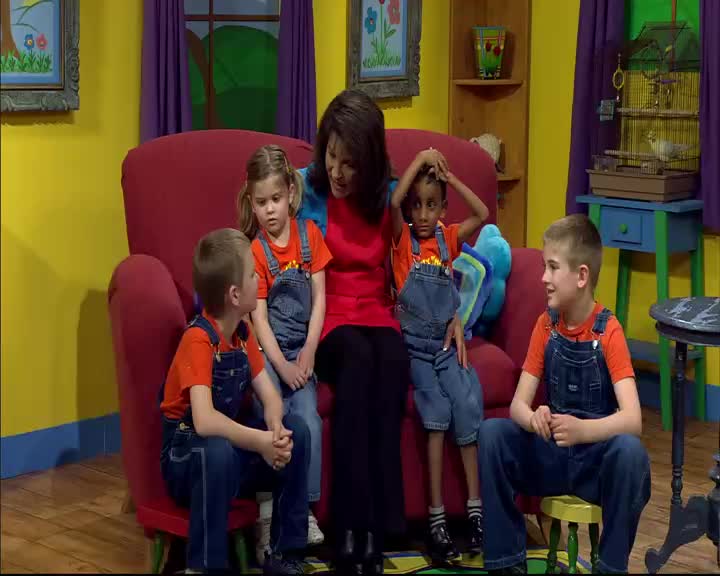 A woman in a red shirt sits on a large red chair, surrounded by four children in orange shirts and denim overalls. Two boys are seated on the floor, one on a small stool, while a girl and another boy sit beside the woman on the chair.