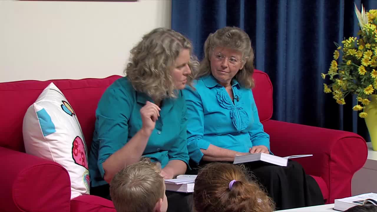 Two women in teal shirts sit on a bright red couch, one gesturing as she speaks. In front of them, children's heads are visible, and a book rests open on the lap of the woman on the right.
