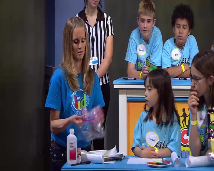 A woman in a blue shirt holds up a plastic bag, preparing for a demonstration. Several children in matching shirts watch intently from behind a table.