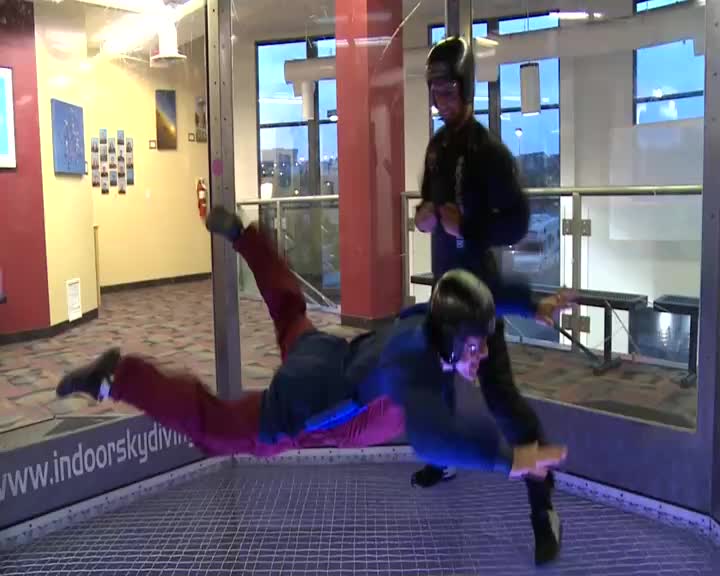 A person in a blue shirt and red pants is suspended mid-air, arms outstretched. An instructor in black stands behind, guiding the flyer.