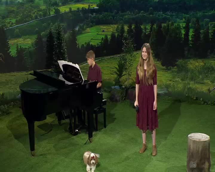 A boy plays the grand piano while a girl stands ready with a microphone. A small dog sits patiently on the green carpet.