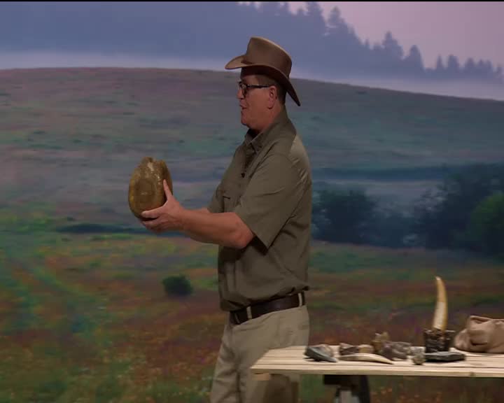 A man in a cowboy hat holds a large, oval-shaped rock. He stands beside a table displaying various fossils and artifacts.