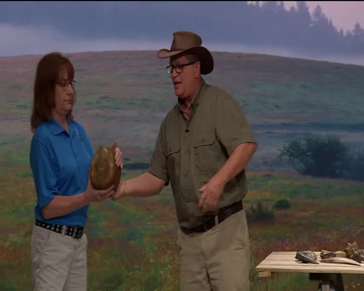 A woman in a blue shirt and a man in a cowboy hat stand on a stage, holding a large, oval object. They appear to be discussing its features, likely for a program like 3ABN Kids.