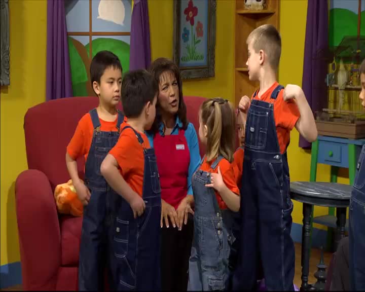 A woman in a red apron speaks to a group of children wearing matching orange shirts and denim overalls. One boy flexes his bicep, showing off his strength.