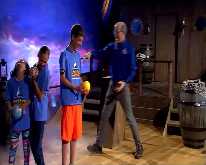 A man in a blue shirt gestures towards a boy holding a yellow ball. Two girls in matching shirts stand nearby, one laughing with a globe in her hand.