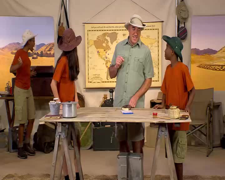A man in a khaki shirt and hat gestures with his finger, explaining something to two children. The children, dressed in orange shirts and khaki shorts, listen intently.