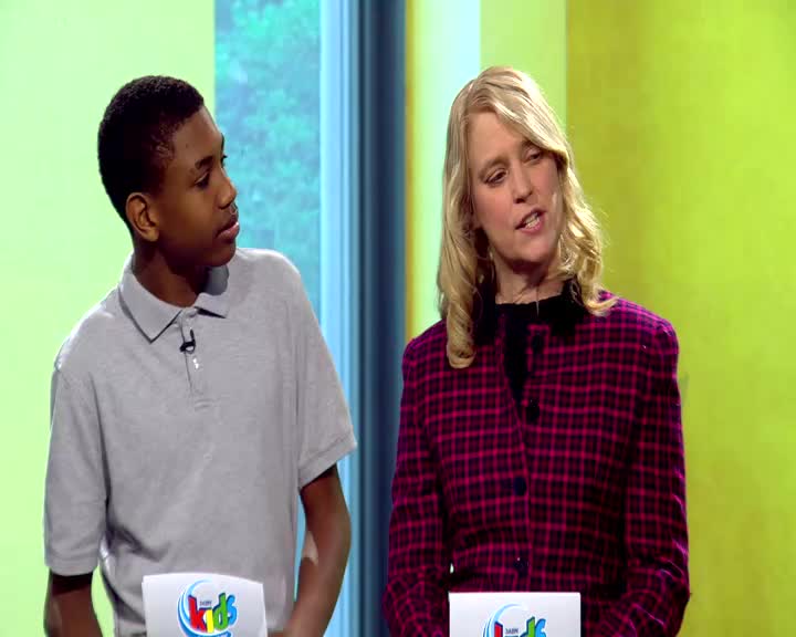 A young man in a gray polo shirt stands next to a woman in a plaid jacket. They are on a brightly lit set, likely for a television program.
