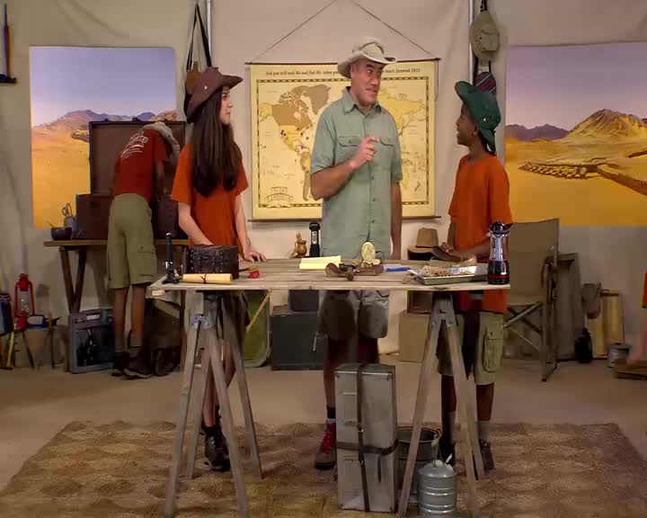 A man in a khaki hat gestures emphatically to a young boy in a green hat, both standing near a table. A girl in a brown hat faces away from them, looking towards a large map on the wall.