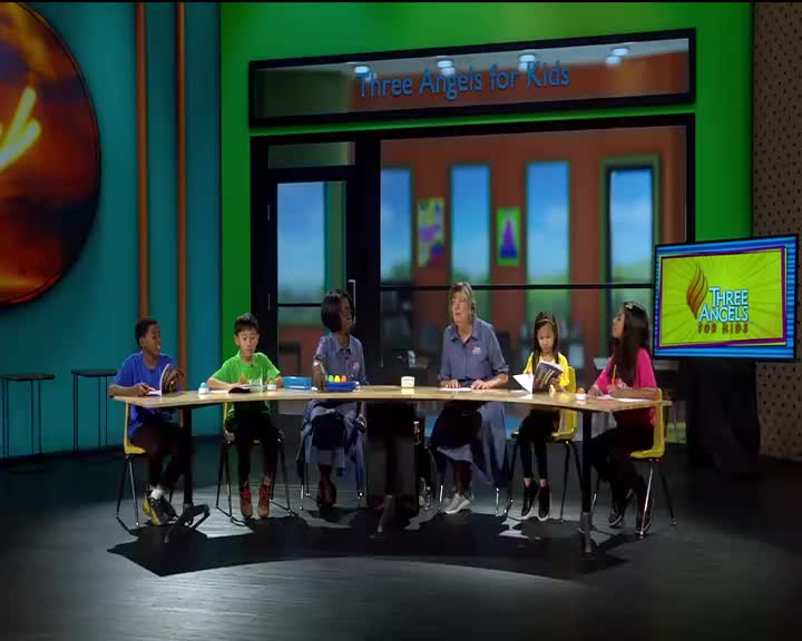 A group of kids sits at a table with a woman, all facing forward. A monitor displays the "Three Angels for Kids" logo.