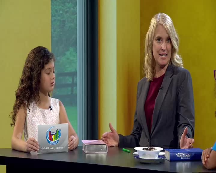 A young girl holds a card with the 3ABN Kids Network logo while a woman in a suit gestures with her hands. A blue book and a bowl of what looks like cereal sit on the table between them.