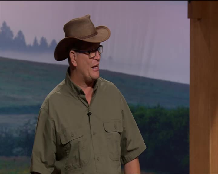 A man in a cowboy hat and green shirt speaks animatedly. He stands before a backdrop of rolling hills.