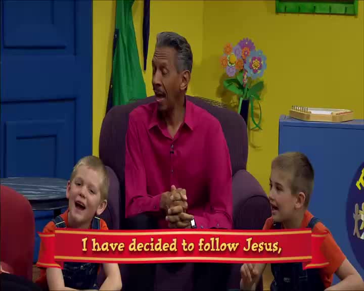 A man in a bright pink shirt sits between two young boys, all looking toward something out of frame. A red banner with gold trim displays the words "I have decided to follow Jesus," as the boys sing along.