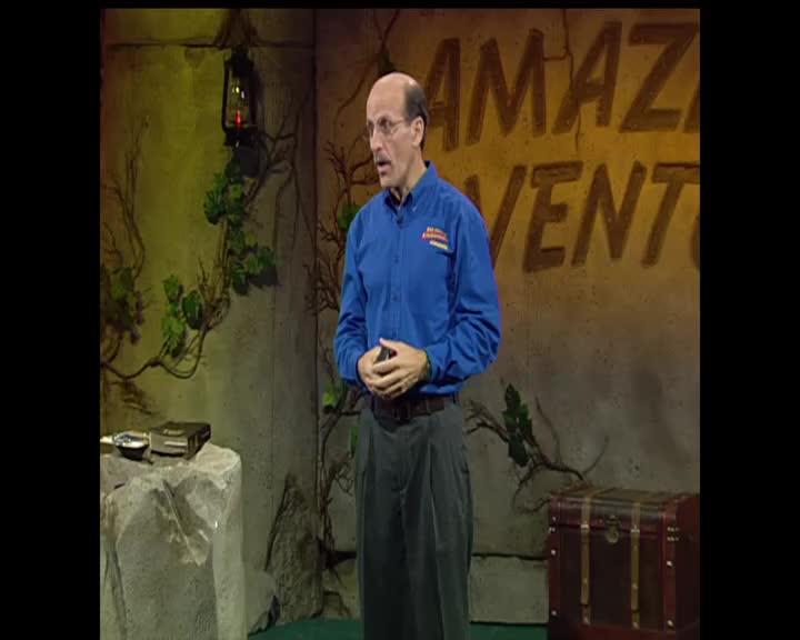 A man in a blue shirt stands before a wall with "AMAZING ADVENTURES" painted on it. He gestures with his hands as he speaks, his gaze directed forward.