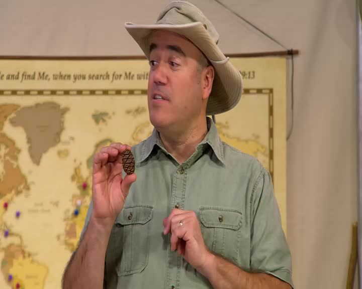 The man in the khaki hat holds up a small, oval object with a diamond pattern. He gestures with his other hand as he speaks.